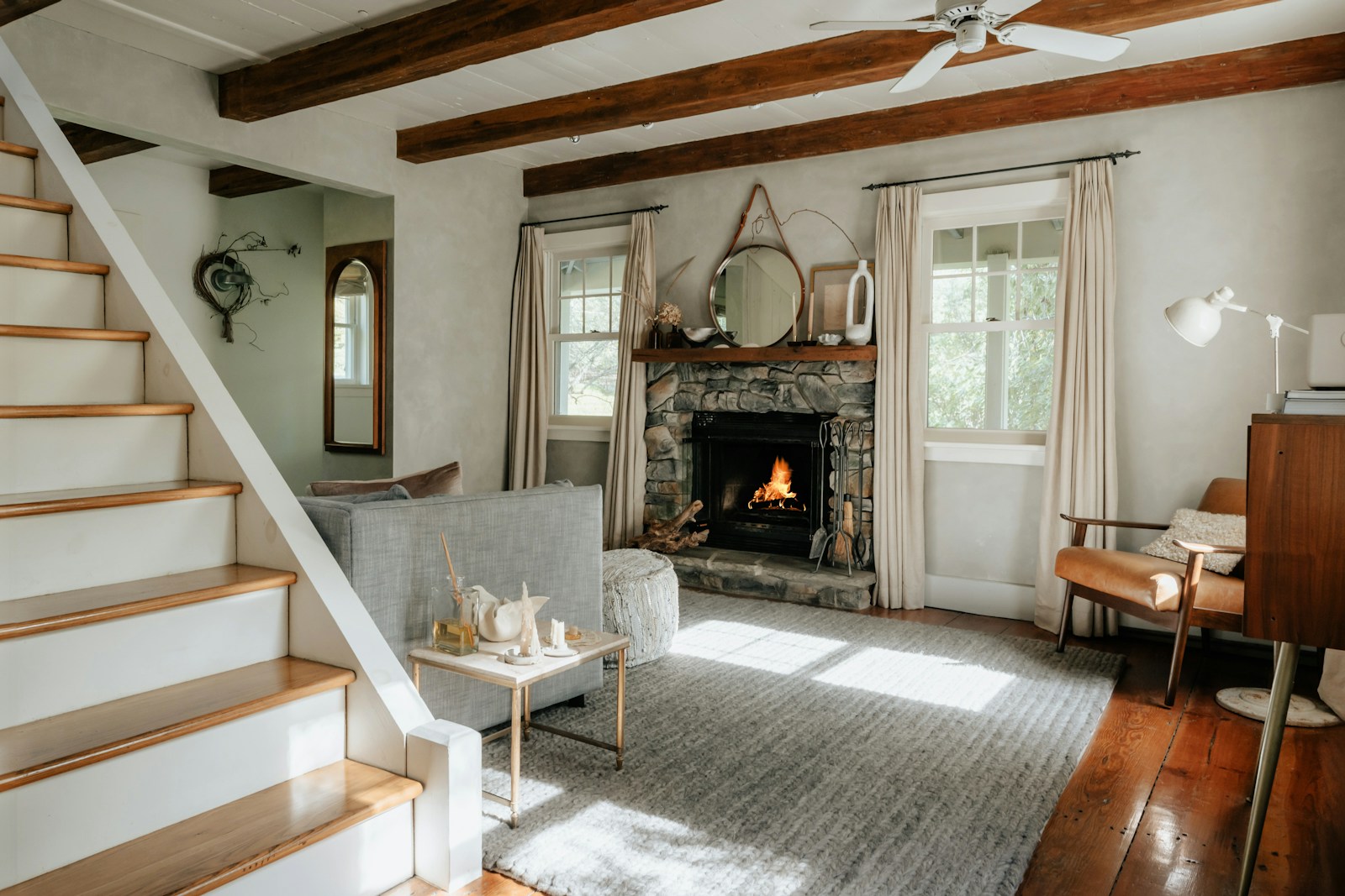 A warm living room with a lit stone fireplace, wood beams and soft daylight through the windows